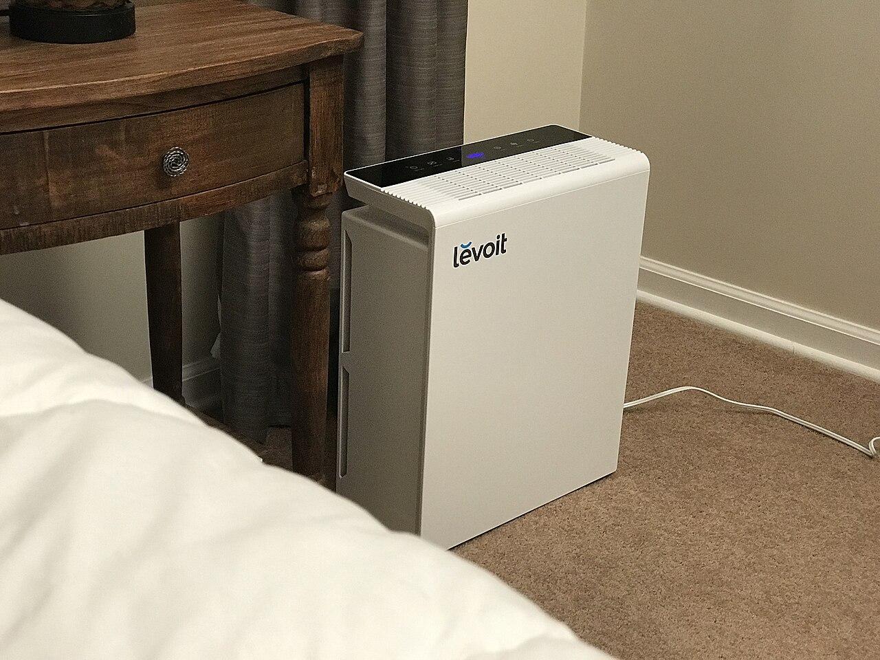 A white air purifier beside a bed in a bedroom corner - three air purifier questions answered